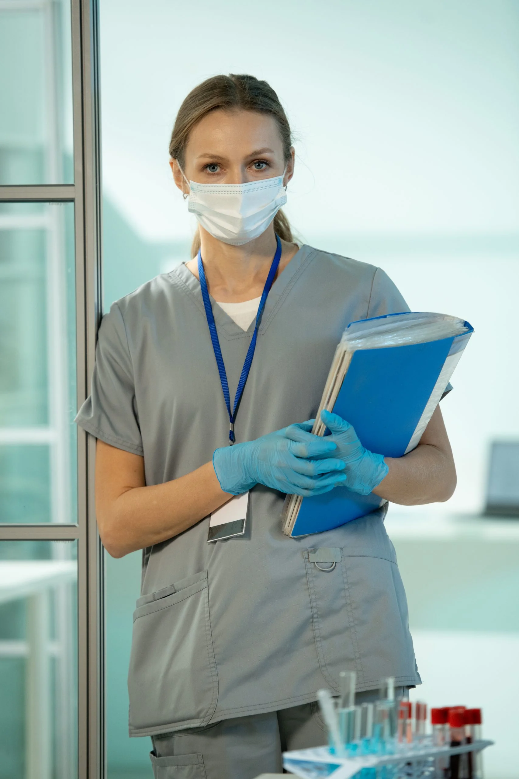 Young serious female clinician in uniform, protective mask and gloves holding medical documents while standing in chemical laboratory